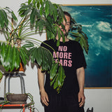 Czarny t-shirt z napisem 'NO MORE TEARS' w różowym kolorze, noszony z czarnymi spodniami, w stylu casual z roślinami w tle