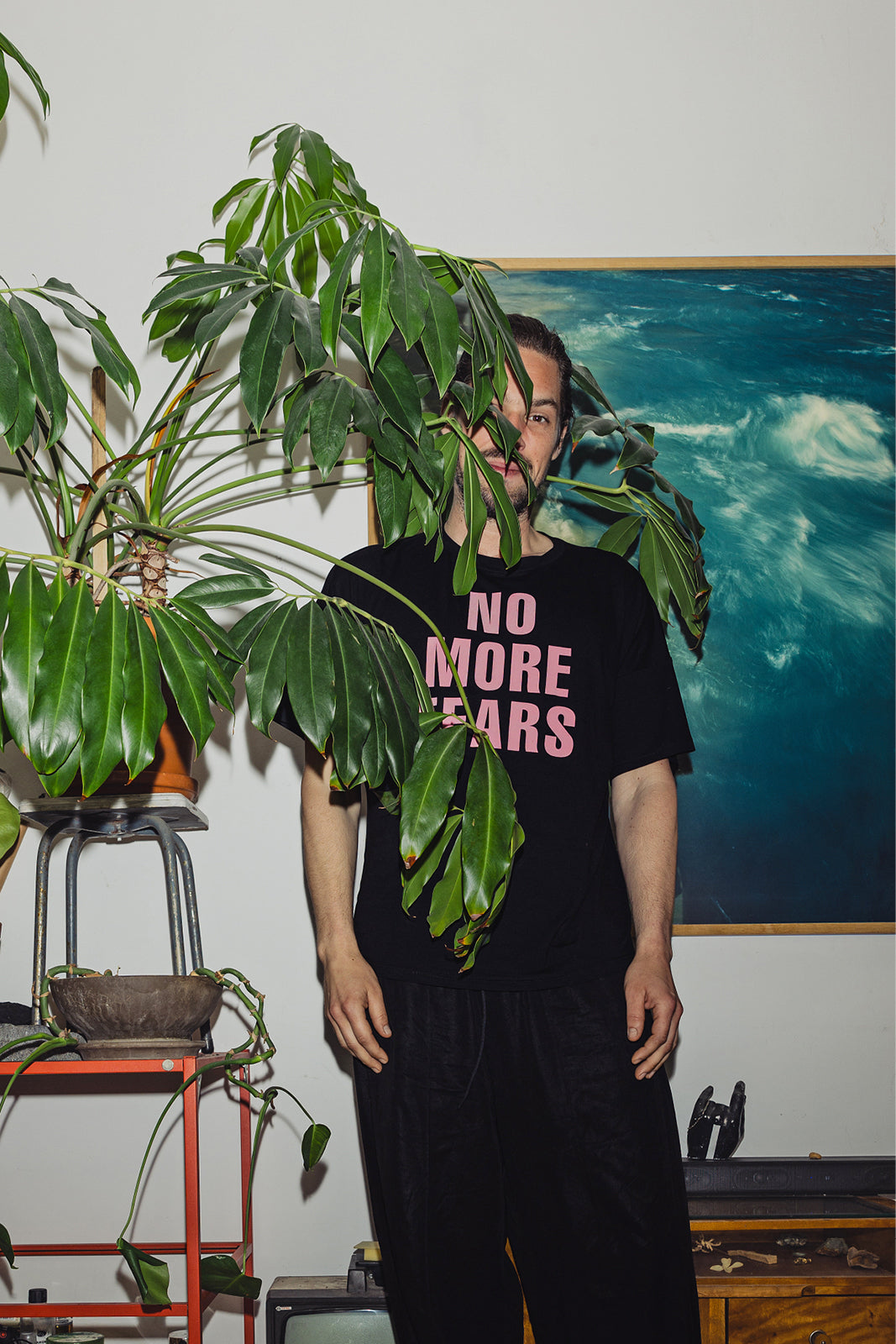 Czarny t-shirt z napisem 'NO MORE TEARS' w różowym kolorze, noszony z czarnymi spodniami, w stylu casual z roślinami w tle