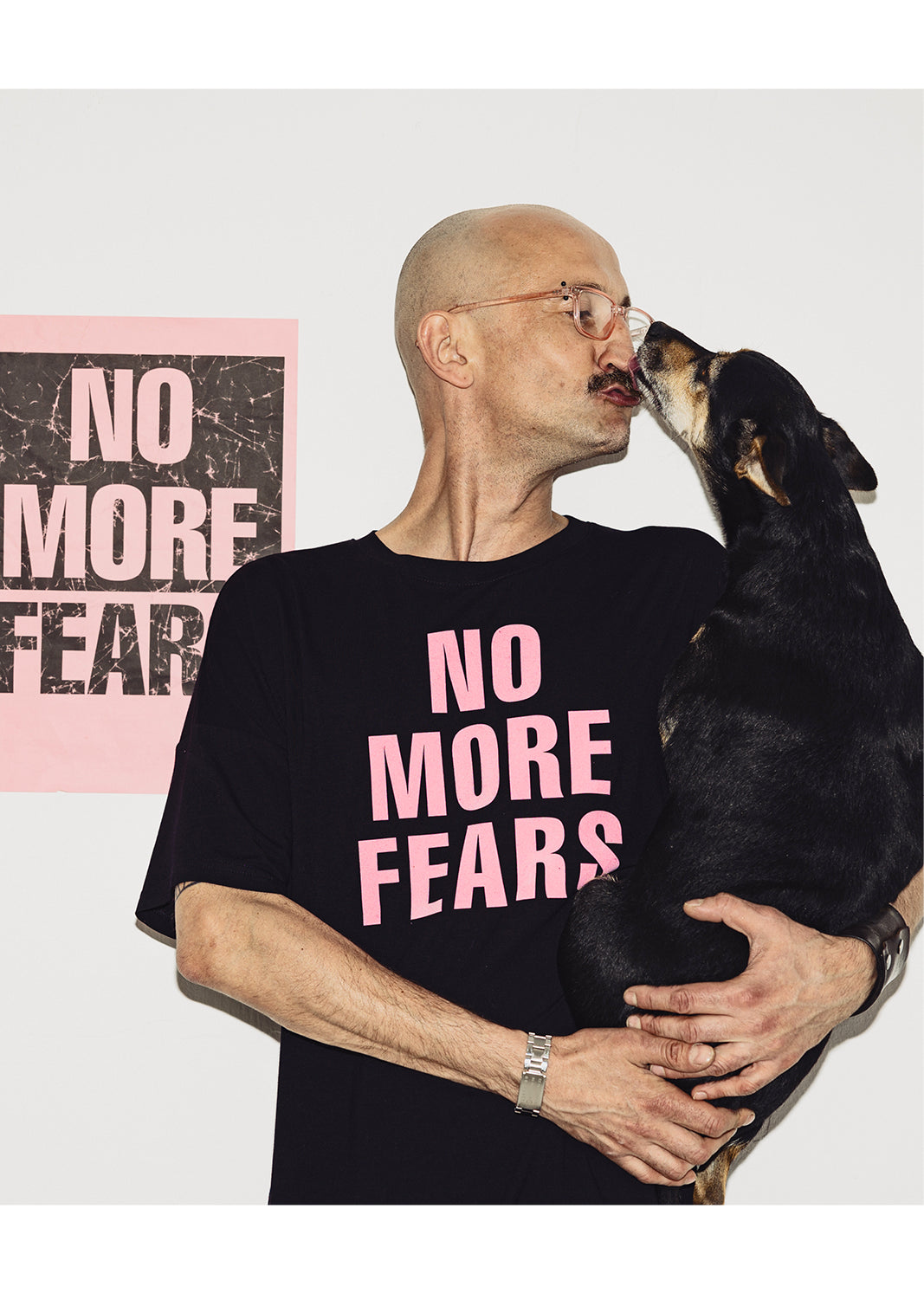 Mężczyzna w czarnej koszulce z napisem NO MORE FEARS przytula psa, tło z plakatem
