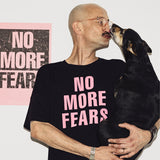Mężczyzna w czarnej koszulce z napisem NO MORE FEARS przytula psa, tło z plakatem