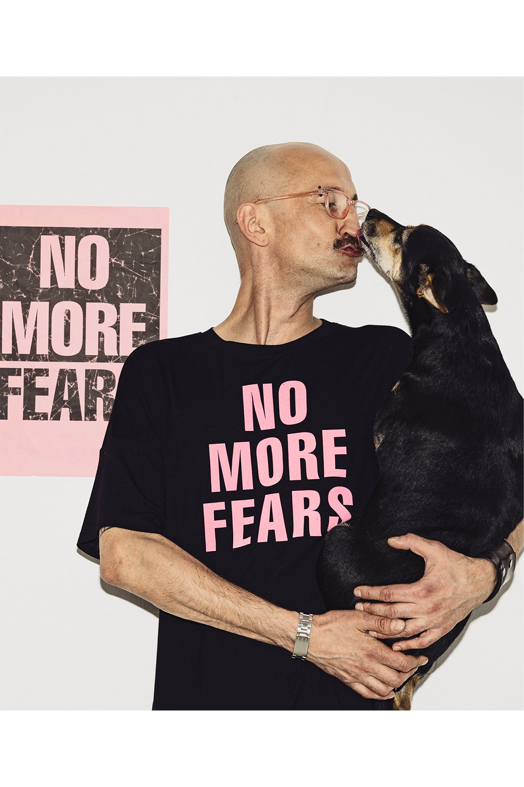 Mężczyzna w czarnej koszulce z napisem NO MORE FEARS przytula psa, tło z plakatem